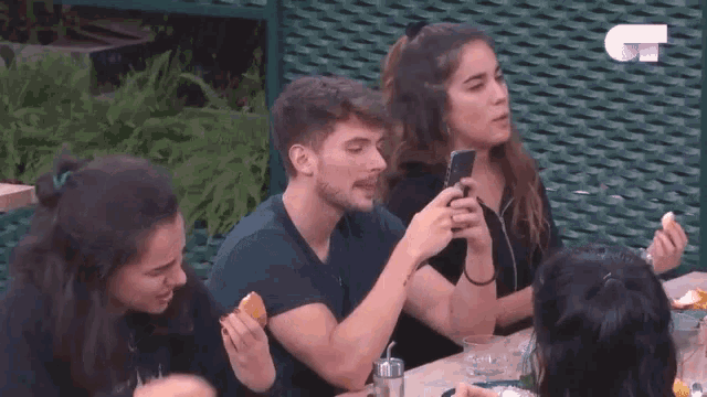 a group of people are sitting at a table eating food and looking at their phones