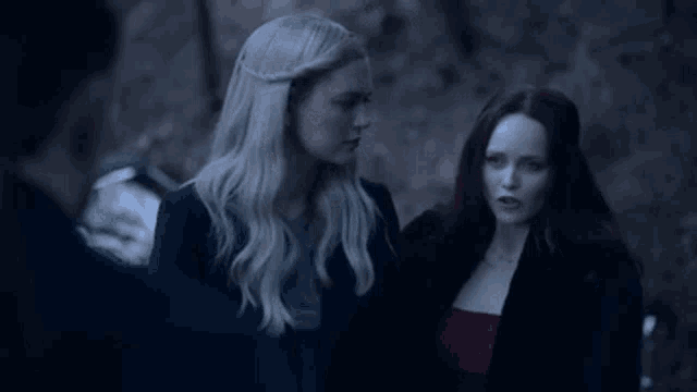 two women are standing next to each other in a dark forest looking at something .