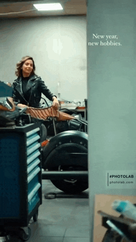a woman in a leather jacket is standing next to a motorcycle in a garage with the words new year new hobbies written on the wall