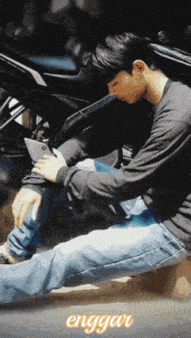 a man is sitting on the ground looking at his phone with the word enggau in the upper right corner