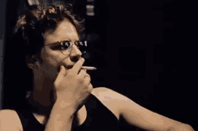 a man wearing glasses and a black tank top is smoking a cigarette in a dark room .