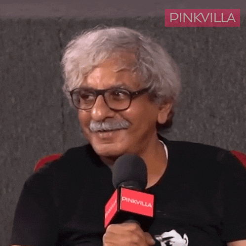 a man wearing glasses and a mustache is talking into a microphone with pinkvilla written on it