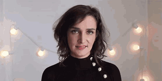 a woman wearing a black sweater with gold buttons is smiling in front of a string of lights