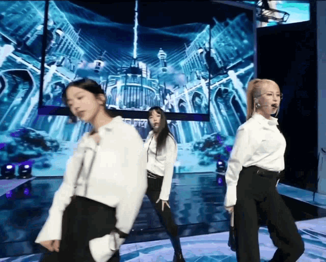 three women in white shirts and black pants are dancing on a stage in front of a screen that says ' a '