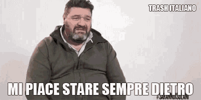 a man with a beard is sitting in front of a white wall and making a funny face .