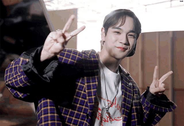 a young man wearing a plaid jacket and a t-shirt giving the peace sign