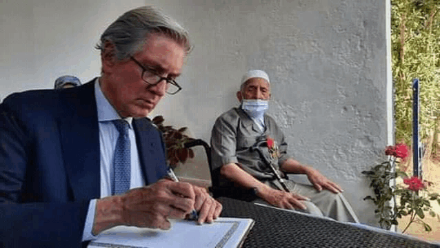 a man in a suit and tie is writing on a piece of paper while another man wearing a mask sits in the background .