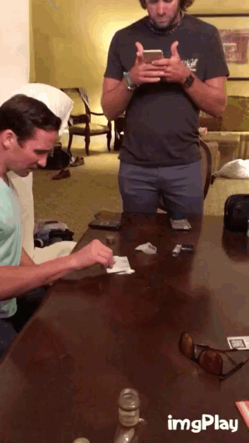 a man sitting at a table looking at his phone while another man sits behind him