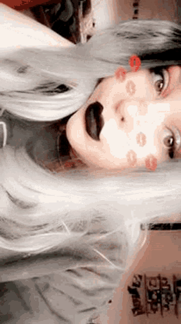 a woman with long gray hair and black lipstick is taking a selfie with kisses on her face .