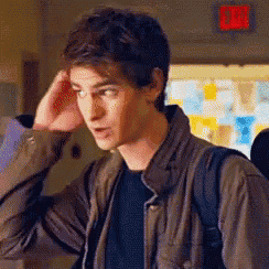 a young man in a brown jacket is standing in a hallway with his hand on his head .
