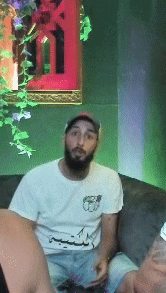 a man with a beard is sitting on a couch wearing a white t-shirt with arabic writing on it