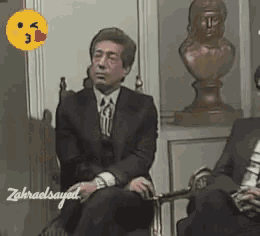 a man in a suit and tie is sitting in a chair with a statue in the background
