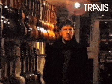a man in a black coat is standing in front of a wall of guitars and the word travis is on the bottom right