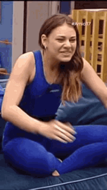 a woman in a blue tank top and blue leggings is sitting on a couch and smiling .