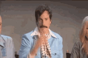 a man with a mustache is wearing a blue jacket and tie while sitting in front of a group of people .