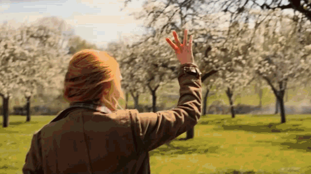 a woman in a brown jacket is standing in a park with her hand up