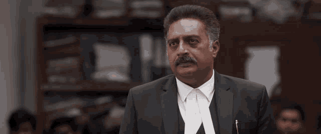 a man with a mustache in a suit stands in front of a crowd