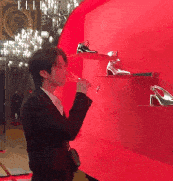 a man in a suit drinking from a glass in front of a red wall that says elle