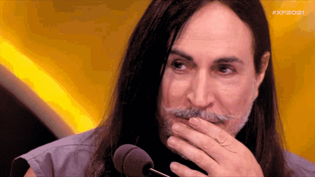 a man with long hair and a mustache holds his hand to his mouth in front of a microphone