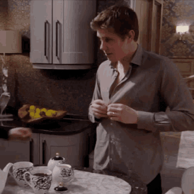 a man in a grey shirt is standing in a kitchen adjusting his shirt