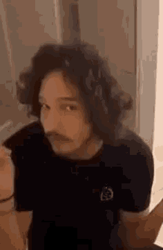 a man with long hair and a mustache is wearing a black t-shirt and looking at the camera .