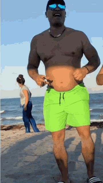 a shirtless man in neon green shorts is standing on a beach