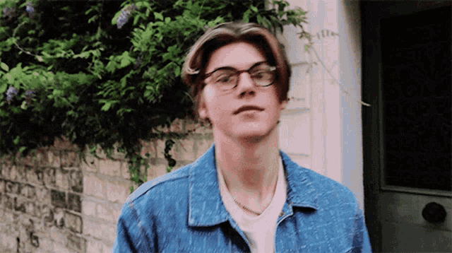 a young man wearing glasses and a denim jacket looks at the camera
