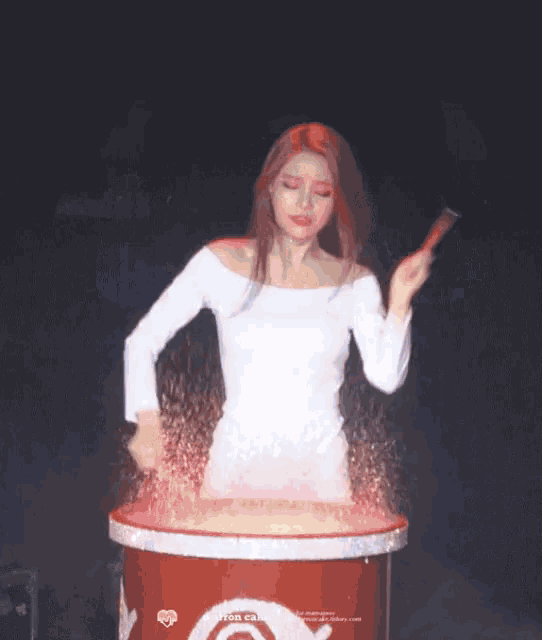 a woman in a white dress is standing in front of a red barrel holding a stick .