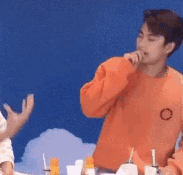 a young man in an orange sweater is sitting at a table with a microphone in his hand .