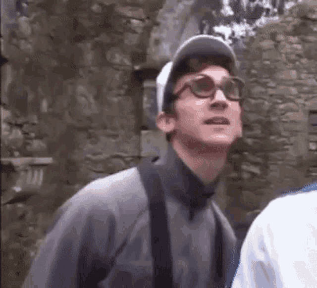 a man wearing glasses and a baseball cap is standing next to a stone wall .