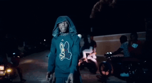 a young man wearing a hoodie and a necklace is standing in a dark street .