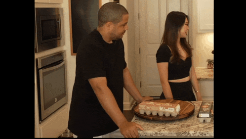 a man in a black shirt stands next to a woman in a black top in front of a tray of eggs
