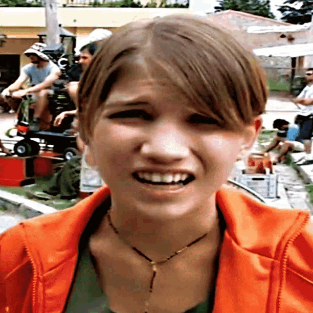 a girl wearing an orange jacket and a necklace is smiling