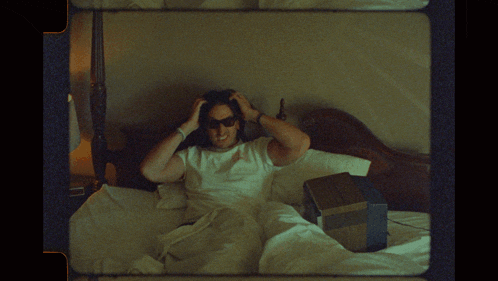 a man wearing sunglasses and a white shirt is laying on a bed with his hands on his head