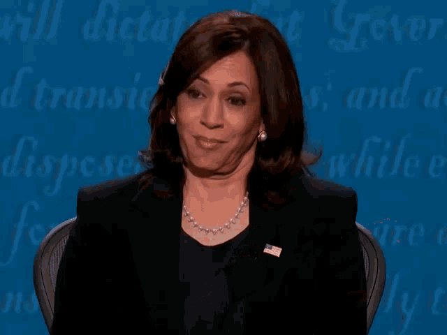 a woman in a black suit and pearl necklace is speaking in front of a blue background that says will