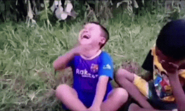 a boy wearing a blue shirt that says rak on it is sitting in the grass laughing .