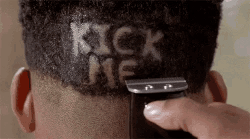 a person is cutting a man 's hair with the words kick me written on his head .