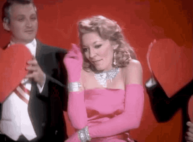 a woman in a pink dress is standing next to a man in a tuxedo holding a red heart .