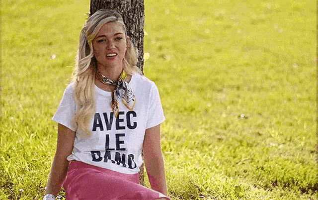 a woman is sitting under a tree in the grass wearing a white t-shirt that says `` avec le band '' .