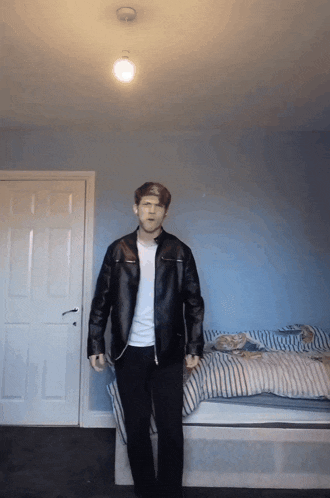 a man in a leather jacket is standing in a bedroom next to a bed