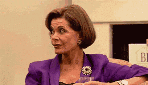 a woman in a purple jacket and brooch is sitting in a chair holding a glass of wine .