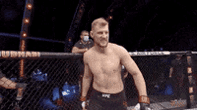 a man without a shirt is standing in a cage with a referee behind him .