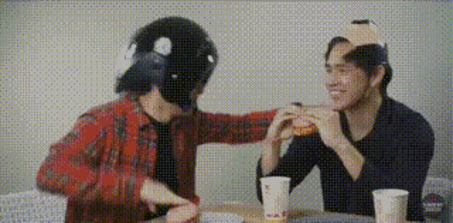 two men are sitting at a table eating hamburgers . one of the men is wearing a helmet .