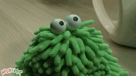 a green stuffed animal with big eyes is sitting on a table next to a coffee cup .