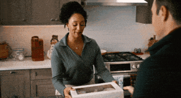 a woman in a kitchen is holding a box of cupcakes and talking to a man