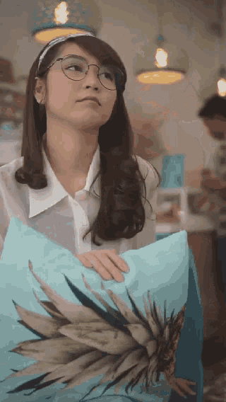 a woman wearing glasses is holding a blue pillow with a pineapple on it