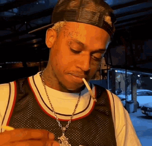 a man with tattoos on his face is smoking a cigarette while wearing a hat .