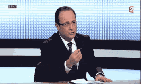 a man in a suit and tie is sitting at a desk in front of a tv screen that says direct