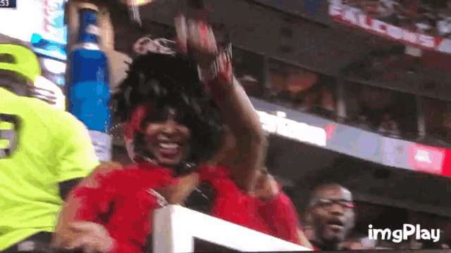 a woman in a red dress is sitting in a stadium watching a game with imgplay written on the bottom right