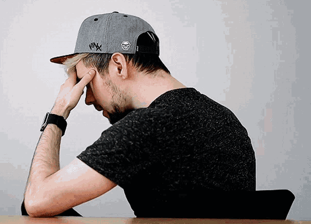 a man wearing a black shirt and a gray hat has his hand on his forehead while sitting at a table